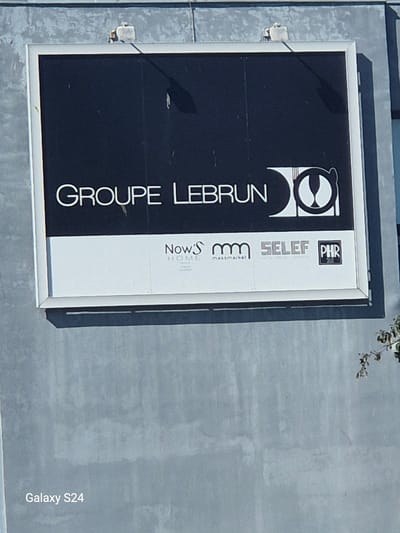 The image shows an aerial view of a large sign on a building. The sign is divided into two main sections. The left section is black with white text that reads "GROUPE LEBRUN". To the right of this text is a logo consisting of two overlapping circles with a white outline.
The right section of the sign is white and contains several smaller logos and text. From top to bottom, these include: a small square logo with the letters "PHR" in white on a black background, followed by the word "SELEF". Below that, there is a logo with the letters "m m" in a stylized font, and below that, the word "massmarket" in smaller text. Finally, at the bottom of this section, is the text "Now's HOME".
The sign is mounted on a building with a gray, textured exterior. Part of a tree with green leaves is visible in the upper right corner of the image. The ground surrounding the building appears to be paved with a gray, concrete-like material. The lighting suggests it is daytime, with shadows cast on the sign. The overall scene is one of commercial signage, likely in an industrial or business park setting. The text "Galaxy S24" is visible vertically along the right edge of the image, indicating it is a captured image from that device.