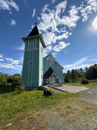 A wide-angle shot captures a light teal-colored church with a tall, slender bell tower against a bright blue sky dotted with fluffy white clouds. The church has a steep-pitched roof and white trim outlining its architectural details. A cross tops the bell tower, and a similar symbol is visible on the side of the church. The church is situated on a grassy hill speckled with small yellow and white wildflowers. A paved patio extends from the church entrance, which features a set of concrete steps. Two dark planters with orange flowers sit on either side of the steps. Beyond the church, a dense line of green trees forms the background. The overall impression is one of a serene, rural setting, with the sun casting a bright light, indicated by a lens flare in the upper right corner. There are no people visible in the image. The location is identified as the village of Läskelä, Russia.