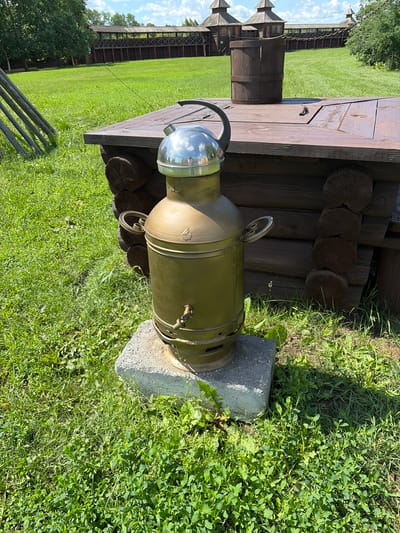 An outdoor shot depicts a brass-colored samovar standing on a concrete base in a grassy area. Behind the samovar, a wooden structure, likely part of a historical settlement or museum, is visible. The structure is made of logs and has a wooden lid. To the left of the samovar, wooden posts form a fence or support structure. In the background, a wooden palisade with watchtowers can be seen, suggesting a historical fortification. The overall setting appears to be a tourist attraction or historical reenactment site. There are no people visible in the image. The sky is partly cloudy.