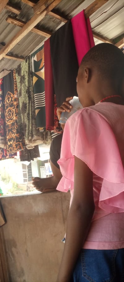 The scene depicts an interior view of what appears to be an open-fronted commercial establishment, likely a fabric stall, in Umuoji, Nigeria. The perspective is from slightly behind and to the right of a primary individual.
In the foreground, a person with short dark hair is observed from the back and right side. This individual wears a light pink, short-sleeved top with layered ruffle detailing on the sleeves and blue denim bottoms. A red beaded necklace is visible around their neck.
To the left and slightly behind the primary individual, a second person is partially visible. This individual holds a clear plastic sachet or container to their mouth, appearing to drink from it. Their bare left foot, with white-painted toenails, rests on a concrete ledge that extends across the lower mid-section of the image, separating the interior space from the exterior. This person wears dark-colored shorts.
Above, the ceiling consists of corrugated metal sheets supported by exposed wooden rafters. From these rafters, multiple rolls of fabric or draped clothing items are suspended. These fabrics exhibit various patterns and colors, including black with gold motifs, green with dark botanical designs, striped patterns, plain dark red, and plain black, suggesting merchandise for sale.
Beyond the concrete ledge, an exterior environment is visible, characterized by soft natural light. Distant structures, including a building with a window frame and hints of vegetation, are present. The wall to the right of the foreground individual is a light brown, possibly unpainted concrete or mud wall, showing some discoloration.