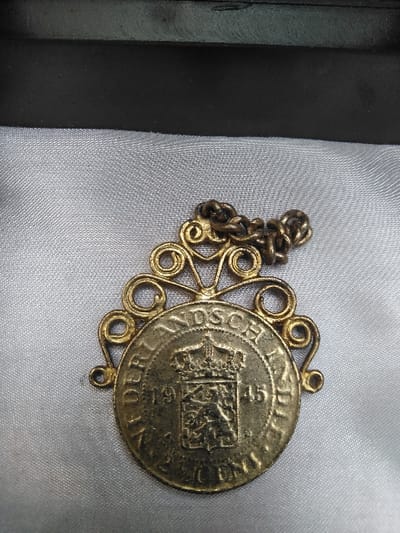 The image presents a close-up, top-down view of a vintage metallic pendant resting on a light grey, satin-like fabric surface. The pendant consists of a circular coin set within an ornate, scrolled filigree frame made of a tarnished golden-colored metal, likely brass or bronze.
The coin, also tarnished and showing wear, displays the inscription "NEDERLANDSCH INDIE" along its upper circumference and "1 CENT" or similar denomination along the lower edge. Centered on the coin's face is a crowned escutcheon featuring a lion rampant, flanked by the dates "19" on the left and "45" on the right, indicating the year 1945. A short, tarnished metal link chain is attached to the uppermost scrollwork of the frame.
The fabric surface beneath the pendant exhibits subtle folds and a slight sheen. In the upper portion of the frame, a dark, rectangular object, possibly the edge of a box or container, is partially visible. No individuals, specific actions, or environmental details that could pinpoint a precise location are discernible within the image. The specific location within Лебяжье ауылдық округі, Kazakhstan, cannot be ascertained from the visual data provided.