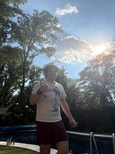 A young person stands outdoors, near what appears to be the edge of a pool, based on the presence of water, pool rails, and bottles near the edge. The individual is wearing a light-colored t-shirt and dark shorts. The person is gesturing with their left hand, possibly making the "shaka" sign, and holding an object in their right hand, possibly sunglasses and a phone. The background shows trees and a bright, slightly cloudy sky with the sun shining. The location is Santa Ana Yancuitlalpan (Chilhuacán), Mexico.