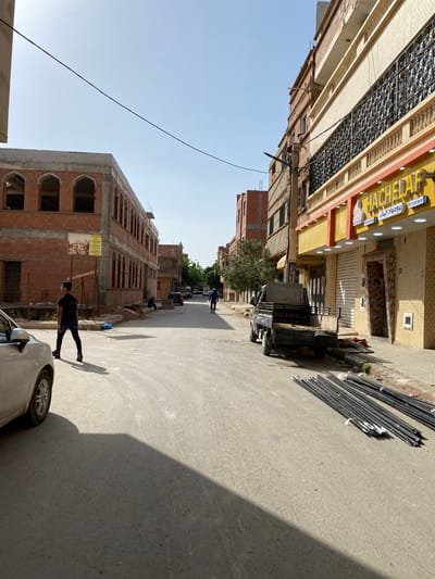 This image captures a vibrant street scene in Sidi Chahmi, Algeria, featuring a mix of commercial and residential buildings under a clear blue sky.

On the left side of the street, a multi-story building with intricate, dark metalwork covering its upper windows or balcony stands prominently. Below it is a storefront with a bright yellow sign displaying Arabic text and two phone numbers: "07 72 01 86 57" and "07 98 83 55 65". Further down the street on the left, a large, multi-story building is under construction, characterized by its red brick facade and numerous arched window openings, with scaffolding and building materials visible around it.

On the right side of the street, there are other buildings with various facades, including one with textured beige walls and another with closed roll-down shutters. A small utility truck is parked here, near a large pile of long, thin metal pipes or rods stacked on the pavement.

Several people are present in the scene. In the immediate foreground, a person in dark clothing and red shoes walks along the street, partially obscured by a silver car whose front section is visible. Another person in blue is walking further down the street, and a person appears to be sitting near the construction site on the left.

The activity depicted is everyday street life, with people moving about and vehicles parked. The construction materials and ongoing work on the brick building suggest development or renovation in the area. Long, sharp shadows cast across the street indicate it is likely midday or early afternoon, with bright sunshine. The visible Arabic text on signs and the architectural styles provide strong location cues for a North African setting.
