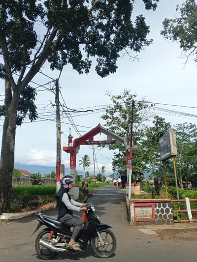 A woman on a motorcycle drives along a road leading to an ornate archway. The archway is painted red and white and is inscribed with the words "MESJID JAMIK AL HIKMAH." The road is lined with green fields and trees. The sky is partly cloudy, and there is a mountain range in the distance. A telephone pole stands to the left of the archway, with many wires running across it.