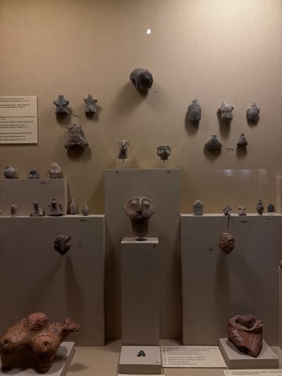 This image captures a museum exhibit within the Archaeological Museum of Drama, Greece. The scene presents a collection of small ancient artifacts, primarily terracotta and clay figurines, busts, and vessel fragments, displayed against a textured beige wall and on light-colored plinths.
On the upper beige wall, numerous small, dark-colored stylized anthropomorphic and zoomorphic figurines are arranged. A rounded dark object is labeled "22" on the left, and two distinct human-like figures are labeled "18-17" in the center. Further right, a cluster of approximately seven additional small figures and objects, including a vessel-like item, are visible, with "38-41" labeling some. Near the top center, a small, dark, mask-like head is mounted on the wall, labeled "23".
Below this wall display, several white or light-colored rectangular plinths support additional artifacts. On the far left, a group of four small, dark, stylized figures is arranged on a plinth, labeled "13-14". Beneath them, a larger, reddish-brown terracotta figurine of a quadruped animal, possibly a bull or similar creature, with prominent features and some surface markings, rests on a lower, wider plinth. Above this animal figure, a dark, circular mask-like object with concentric patterns hangs on the wall, labeled "3".
In the center of the image, a tall, white rectangular plinth supports a light-brown terracotta bust, depicting a human head with large eyes and an elongated face, labeled "19". To its left, a smaller, dark, stylized head on a narrow stand is labeled "21".
On the right side, multiple small, dark, stylized figurines are displayed on an elevated plinth, with the group labeled "26-29". Below this, a reddish-brown, mask-like facial fragment hangs on a smaller plinth, labeled "24". Further right, on a wide, low white plinth, a large, reddish-brown ceramic vessel fragment or stylized bust with vertical painted stripes of a darker hue is displayed, labeled "23" at its base.
An informational text panel, partially visible on the far left, provides descriptions in Greek and English for various categories of figurines, including "domestic animals," "violin-shaped figurines," "seated female figurines," "pregnant female figurines," and "miniature clay masks," with phase designations (Phases I, II, III). Specific labels on the plinths, such as "19 (Masked?) head of a male figure (Phase III)" and "22 Miniature clay mask (Phase III)," correlate with the text panel's content.
The environment is illuminated by artificial lighting, creating shadows beneath the wall-mounted objects and highlighting the displays. The overall impression is that of a professional museum exhibition showcasing ancient archaeological finds.