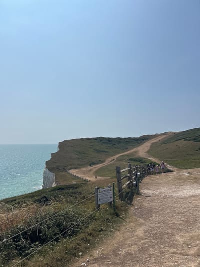 The image shows a section of the Seven Sisters cliffs near Seaford, United Kingdom. A dirt path winds along the cliff edge, with a wooden fence bordering it. Several people are walking on the path. The sea is to the left of the frame, and rolling green hills stretch into the distance. A "DO NOT PASS" sign is visible on a post near the fence, warning of the dangers of chalk cliff falls and unstable edges. The sky is clear and blue.