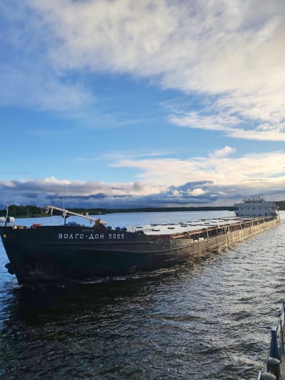 A wide-angle shot depicts a cargo barge on a river under a partially cloudy sky. The barge, with the name "ВОЛГО-ДОН 5065" painted on its bow, is dark in color and appears to be of older construction. It is moving through the water, which has a dark blue-green hue. The sky features a mix of blue and white clouds. The far shore is visible as a line of trees in the distance. The scene is a river landscape, possibly near a town or rural area. The presence of the barge suggests industrial or transportational activity.
There is no information available about the location in the city of Pochinok, Russia.