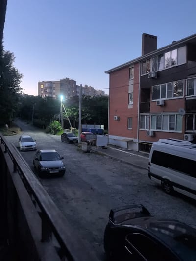 The image shows an outdoor scene at dusk, likely in a residential area of Verkhnetamarlytsky, Russia. A multi-story brick apartment building is prominently featured on the right, with windows reflecting the twilight sky. A white van is parked in front of the building. To the left of the building, a gravel road slopes uphill, lined with trees and other apartment buildings in the distance. Several cars are parked on the road, including a silver sedan in the foreground. A street light is illuminated on a pole near the center of the road. The overall impression is of a quiet evening in a suburban or semi-urban environment.