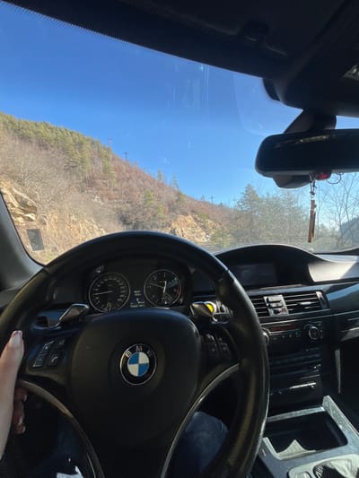 The image depicts the interior of a BMW vehicle from the driver's perspective, located in Erma Reka, Bulgaria. A left hand, featuring painted fingernails, is positioned on the black leather steering wheel. The vehicle's instrument cluster displays a time of 09:57, an ambient temperature of +3.5°C, and a current speed of 24 km/h. A wooden cross is suspended from the rearview mirror. Through the windshield, a clear blue sky is visible above a mountainous or hilly landscape. The terrain features steep slopes covered with a mix of deciduous trees (currently bare) and coniferous trees. A dirt or rocky embankment is visible immediately to the left of the apparent road. Multiple utility poles, equipped with crossarms, are situated on the hillsides, indicating the presence of power lines.