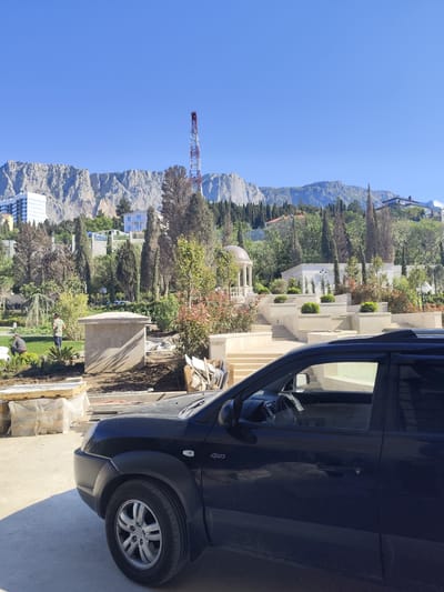 The image shows a black car parked in front of a construction site. In the background, there's a terraced garden with a gazebo and a mountain range. A red and white radio tower can be seen on one of the mountains. There are two people in the background. The location is likely near a coastal area with mountainous terrain and modern buildings. The image does not appear to be in George, South Africa.