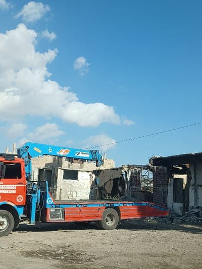In Al-Qadam Municipality, Syria, a red and blue flatbed tow truck with an attached blue TRIANNO hydraulic crane is parked on unpaved ground. The truck's cab displays partial contact numbers, "5958656064" and "998553895," along with a circular emblem featuring a figure in profile. Behind the vehicle, two severely damaged buildings are visible. The structure directly behind the crane is partially collapsed, with exposed concrete blocks and internal debris. The adjacent building to the right exhibits significant fire damage, characterized by a blackened interior, collapsed roof sections, and structural disintegration. Extensive rubble and debris are scattered across the ground in front of the damaged structures. The sky overhead is clear blue with scattered white cumulus clouds. No individuals or active interactions are present in the scene.