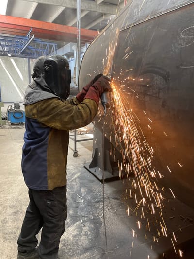 A welder works on a large metal structure in a brightly lit industrial setting. The welder, wearing a protective mask, gloves, and jacket, holds a grinder that emits a shower of orange sparks. The metal structure appears to be part of a large cylindrical object supported by metal stands. The floor of the facility is light gray. Other structural elements of the building are visible in the background, including a large red support beam and a blue scaffolding structure. The presence of welding equipment nearby indicates this is a fabrication or repair operation. There is no conclusive evidence to verify if the location is actually in Zaljevo, Montenegro.
