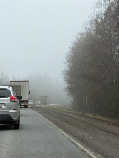 The image shows a traffic jam on a road in Bolgury, Russia. The scene is dominated by heavy fog. The road is wet, and a white line marks the edge. A silver minivan is visible in the foreground, with other vehicles, including a truck, lined up behind it. Bare trees are visible along the roadside, adding to the stark atmosphere. The overall impression is one of poor visibility and disrupted traffic flow.
