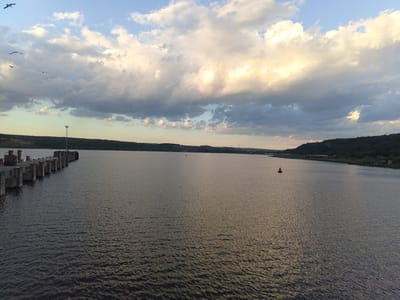 The image shows a wide body of water, likely the Beloslav Lake in Bulgaria, based on similar landscape features. A pier or dock extends from the left edge of the frame into the water. A single red buoy is visible in the middle of the water. The shoreline in the distance features a hilly landscape with sparse vegetation. The sky is filled with clouds, ranging from light and bright to darker and more ominous. There are some birds flying in the sky on the left.