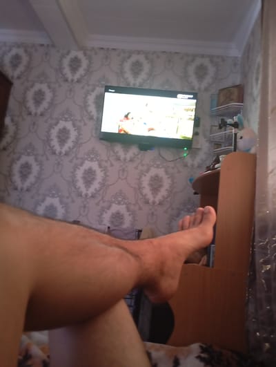 In a dimly lit room with patterned wallpaper, a person's bare legs and feet are prominently featured in the foreground, suggesting they are resting or relaxing. A television screen displays an image of people, possibly in an entertainment or news program. To the right, a wooden shelf unit holds various items, including a plush toy, and a bedside table is visible. The overall impression is of a casual domestic setting, likely a bedroom or living room. The city and country mentioned in the prompt are not visually identifiable from the image.