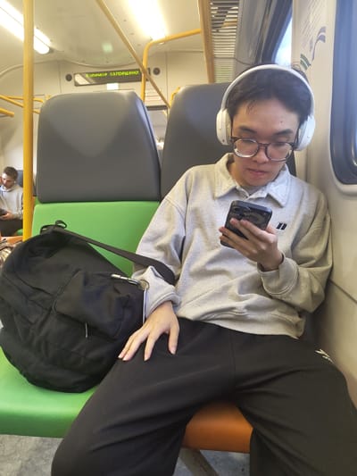 A young person wearing headphones and glasses is sitting on a train and looking at their phone. They are wearing a light grey sweatshirt and black pants. A black backpack is on the seat next to them. In the background, another person is sitting on the train and looking at their phone. The train appears to be in motion, with yellow handrails and grey seats visible. The text on a digital display above the seats reads "Курение запрещено!" which translates to "Smoking is prohibited!" in Russian. The scene takes place inside a train car, likely within the city of Nizhnee Khoroshovo, Russia.