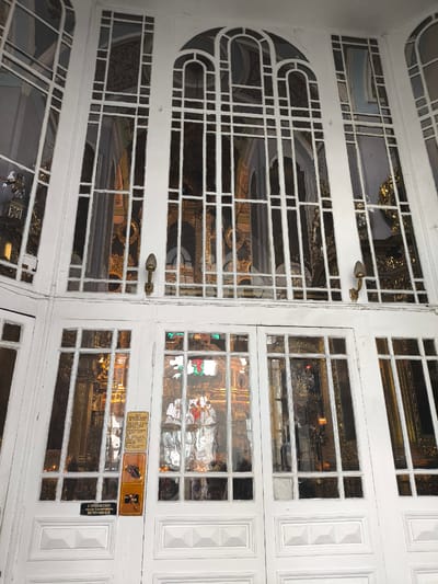 The image captures the grand, white-painted entrance of a religious building, likely a church or cathedral, viewed from the exterior during the daytime. The entrance features an intricate design with numerous glass panels set within white wooden or metal frames, including large arched sections at the top and rectangular panes below.

Through the glass, the opulent interior of the building is visible, characterized by golden decorations, religious iconography, and possibly an iconostasis or altar. A red light source, perhaps a lamp or candle, can be seen within the reflections. Stained glass elements are also visible in the central lower door panels. Two brass wall-mounted light fixtures (sconces) are attached to the exterior frame above the doors.

Several signs are affixed to the central lower door. The top yellowish-brown sign, titled "ДЛЯ ПОСЕТИТЕЛЕЙ ХРАМА" (For visitors of the temple/church) in Russian, lists rules for visitors, mentioning aspects like removing headwear, lighting candles, and prohibitions against smoking. Below this, two orange signs with symbols are present: one appears to prohibit a bag or certain items, and the other clearly depicts a camera with a diagonal line through it, signifying "No Photography." A black sign at the very bottom further reinforces this rule with the Russian text: "В ПОМЕЩЕНИИ СОБОРА ФОТО И ВИДЕОСЪЁМКА ЗАПРЕЩЕНЫ" (Inside the cathedral, photo and video shooting are prohibited). The architectural style and Russian text are consistent with a sacred site in Oktyabrskoye, Russia. No people are visible.