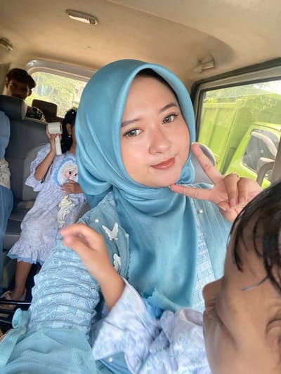 The image captures the interior of a vehicle in Ketegan, Indonesia. A light-skinned woman, wearing a light blue hijab and a matching top embellished with white butterflies and pearl beads, is prominently featured in the foreground, looking directly at the camera with a subtle smile and displaying a peace sign with her right hand. A small child's dark-haired head is partially visible in the bottom right corner. In the midground, a second child, wearing a light-colored dress with a blue pattern and an Elsa character graphic, is seated and holding a roll of white toilet paper in her right hand. Behind this child, a man with dark hair is partially visible. The vehicle's off-white ceiling is discernible, and through a window in the background, bright green foliage and a portion of a bright green truck or utility vehicle are visible outdoors.
