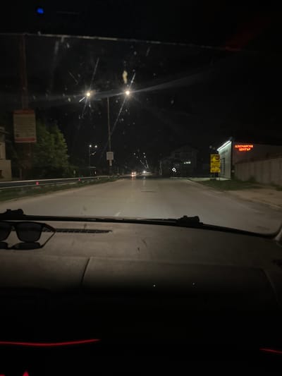 The image shows a night-time view from inside a vehicle, looking out onto a street in Yasen, Bulgaria. The perspective is from the driver's seat, with the windshield and dashboard visible. The road ahead is illuminated by streetlights and the headlights of other vehicles. Trees and buildings line the street, and a few signs are visible. The overall mood is somewhat eerie due to the darkness and the low quality of the image.
One prominent object in the scene is a pair of sunglasses resting on the dashboard. On the left side of the image, a building with a bright red neon sign is visible. The sign displays text in Cyrillic script, which appears to read "КЛЮЧАРСКИ ЦЕНТЪР," meaning "Locksmith Center."
The time of day is clearly night, as indicated by the darkness and the artificial lighting. The weather appears to be clear, as there is no visible precipitation or fog. The scene depicts a typical street at night, with vehicles moving along the road and businesses illuminated. The absence of people suggests that it is late at night or early in the morning. The image is slightly blurry and grainy, possibly due to low light conditions or the quality of the camera used.