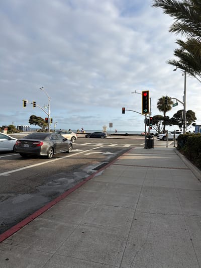 The image shows a street scene in Laguna Beach, California, USA. A traffic light is visible, displaying a red light and a green arrow. Several cars are present on the road, and pedestrians are walking on the sidewalk. The background features the ocean and palm trees, indicating a coastal location. The weather appears to be partly cloudy.