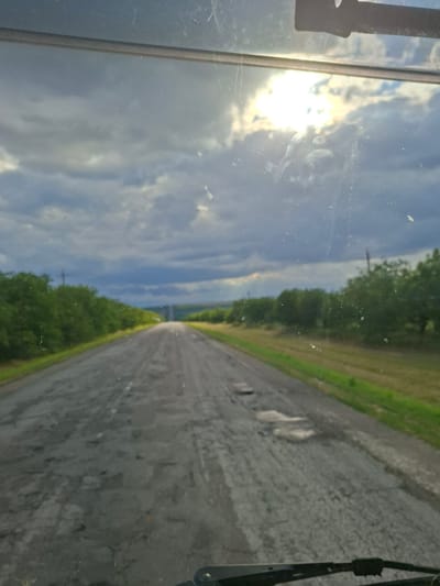 The image shows a damaged asphalt road stretching into the distance, lined with green trees and fields. The sky is cloudy, with the sun breaking through in some areas. The view appears to be from inside a vehicle, looking out through a windshield that has some smudges and dirt on it. There are no people visible in the image. The road has numerous potholes and uneven surfaces, indicating poor condition. The overall impression is of a rural or semi-rural landscape. Without additional information, it is not possible to confirm the specific location as Florești, Moldova.