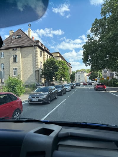 The image captures a traffic jam on a street in Giessen, Germany. The perspective is from inside a vehicle, looking out towards the right. A line of cars is visible, stretching down the road, indicating slow-moving or stopped traffic. The cars are of various colors, including dark gray, black, and a red one in the foreground.

The scene is set on a paved road with lane markings. On the left side of the road, there are buildings with brick facades and multiple windows, typical of a residential or mixed-use area. Trees with lush green foliage line the street and are interspersed between the buildings. The sky is blue with scattered white clouds, suggesting fair weather during the daytime. The sunlight casts shadows, indicating it is likely midday or afternoon.

Some visible text includes license plates on the cars, such as "KUS N 793" on a dark gray car. There are also street signs with German text and arrows, one of which appears to show speed limits. The overall impression is a typical urban scene experiencing traffic congestion. There are no people clearly visible within the frame.