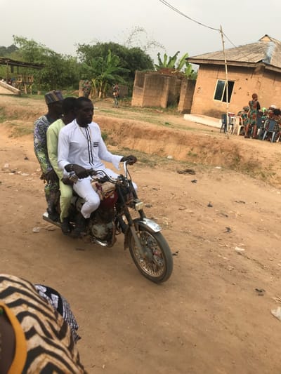 Three individuals are on a motorcycle. The driver, in a white traditional outfit, is focused ahead. Behind him are two passengers. They are on a dusty, unpaved road with a building and vegetation visible in the background. The setting appears to be a rural area. There's a group of people gathered in front of the building. A person wearing an animal print is also visible in the right bottom of the picture.