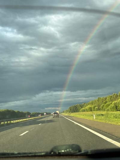 The image captures a view from a vehicle driving on a highway. The scene is dominated by a cloudy sky with a visible rainbow arching across it. To the right of the road, a dense line of green trees runs alongside. The highway itself is paved and has white lane markings. Several vehicles, including cars and trucks, are visible on the road ahead. The lighting suggests it might be daytime, possibly after a rain shower, given the presence of the rainbow and the overcast sky. The overall mood is serene, perhaps tinged with awe at the natural phenomenon. No visible text is present in the image.