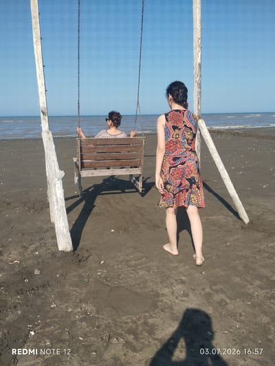 In Abilyatag, Azerbaijan, a sandy beach scene unfolds with two women positioned near a wooden swing set. One woman, seated on the swing, faces away from the camera, her hair pulled back and wearing sunglasses. The second woman stands in front of the swing, looking out at the sea. She is wearing a colorful, patterned dress. The environment is characterized by a dark sandy beach, the calm sea, and a clear blue sky. The swing set appears weathered, constructed from rough wooden posts and metal chains. The lighting suggests a sunny day.