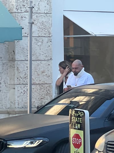 A man and a woman stand near a black car with a "State Law - Stop for" sign on the door frame. The man is bald and wears a white shirt. The woman has dark hair and her hand is near her head. They are partially obstructed by the car. In the background, there is a light-colored building with a geometric window design and a textured wall.