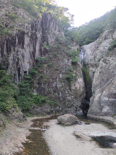 A vertical photograph captures a rugged natural gorge located in Godang-ri, South Korea. The scene is dominated by steep, rocky cliffs on both sides, with a narrow waterfall and a shallow riverbed at its base.

The left side of the frame features a dark, weathered rock face exhibiting prominent vertical striations and patches of green moss and sparse vegetation. The cliff rises sharply towards the top, where dense green foliage is visible against a bright, hazy sky.

The right side presents a lighter-toned, rugged rock face, also covered with green vegetation. A narrow waterfall descends from the upper-right portion of this cliff, cascading into a shadowed, crevice-like pool at its base. More dense green trees line the top edge of this cliff against the bright sky.

At the bottom of the gorge, a wide, shallow riverbed is composed of light-colored sand, gravel, and scattered pebbles. A clear, shallow stream flows through the center of this bed, reflecting light from the sky. Darker, possibly stagnant, pools of water are present in the lower right.

A large, irregular, greyish-brown boulder is situated prominently on the sandy bank in the lower-center-right of the image. Adjacent to this boulder, a single individual is observed in a seated or crouching position, dressed in dark clothing, facing towards the water and the waterfall. The individual's activity is not explicitly discernible.

The overall environment is natural and rugged, illuminated by bright daylight, which highlights the upper sections of the cliffs and foliage.