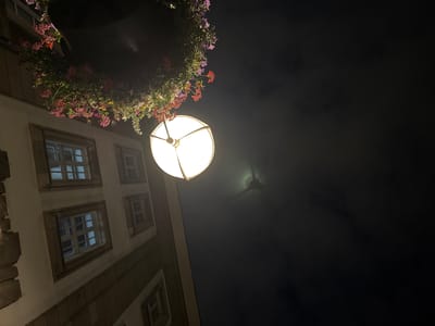 The image, taken at night, shows a building facade with multiple windows and a hanging flower basket. A street lamp illuminates the scene. In the distance, a faint outline of a structure can be seen through clouds, possibly a landmark in Duisburg, Germany. The perspective is from below, looking upwards.