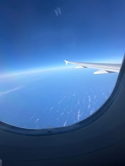 The image is taken from inside an airplane window, looking out at a clear blue sky with a few wispy clouds below. An airplane wing, painted white, juts into the frame from the left, extending diagonally upwards. The wing has multiple sections and appears to be in good condition. The curvature of the Earth is visible at the horizon, with a subtle gradient of blue from deep indigo at the bottom to a lighter, brighter blue at the top, suggesting a high altitude. The sun's glare is prominent on the left side of the image, creating a bright halo effect. The window frame, dark gray and rounded, partially obscures the view on the right side. The overall scene conveys a sense of travel and perspective from above the clouds. The image was likely taken during the daytime, given the bright sunlight and clear skies.