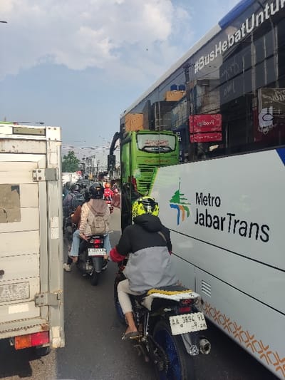 The image shows a scene in Kecamatan Cimahi Selatan, Indonesia (latitude: -6.899, longitude: 107.562). It is a narrow, congested street with a large white bus parked on the right side of the road, partially blocking the road. The bus is a Metro JabatTrans with the words "#BusHebatUntuk" on the side and "Metro JabatTrans" in red on the white body. The bus is facing the right side of the image and is parked with its back end facing the camera. The front of the bus is partially obscured by a smaller green bus behind it. The front of the white bus reflects other buildings. There are three motorcycles on the left side of the image, all in the middle of the road, moving toward the camera. The person on the front motorcycle is wearing a grey hoodie and beige pants. The person on the middle motorcycle is wearing a beige cardigan over a blue shirt and black pants. The person on the back motorcycle is obscured by the vehicle. All three motorcycles are traveling in a single lane. The scene takes place on a heavily trafficked street with several vehicles on the road and buildings and trees in the background. The sky is mostly cloudy. The overall scene is indicative of a traffic jam in a busy city environment.