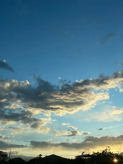 The image shows a sky with a clear blue expanse on the left, transitioning to areas filled with clouds towards the right. The clouds vary in color from white and gray to darker, more ominous shades. On the far right, silhouetted trees and a building are visible, indicating the edge of a town or settlement. The lighting suggests either sunrise or sunset, with the sun positioned behind the trees, creating a bright glow. The image was likely taken outdoors in Khoronq, Armenia.