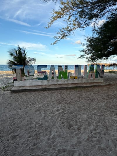 The image depicts a static scene on the San Juan beach in Tela, Honduras. In the foreground, sandy terrain with visible footprints occupies the lower portion of the frame. Centered horizontally in the midground is a large, colorful sign spelling "I ❤️ SAN JUAN". The individual letters are robust, possibly constructed from concrete or a similar material, and are painted with distinct scenes and patterns. The 'I' is predominantly red. The heart symbol replacing the 'O' contains a depiction of a robed figure holding a staff, set against a blue and white background. The subsequent letters, "S", "A", "N", "J", "U", "A", "N", feature various shades of blue, green, and yellow, illustrating landscapes or structures. This sign rests on a grey concrete base. To the left of the sign, a green palm tree is partially visible. To the right, a darker, pine-like tree extends into the upper right quadrant of the image. A single individual is partially visible behind the sign, positioned between the 'N' and 'J' of "SAN JUAN", with only their lower torso and legs, clad in dark attire, discernible. The background features the calm, blue ocean with a discernible horizon line, under a light blue sky with sparse, wispy white clouds. On the far right, a structure with a thatched roof is visible on the beach.