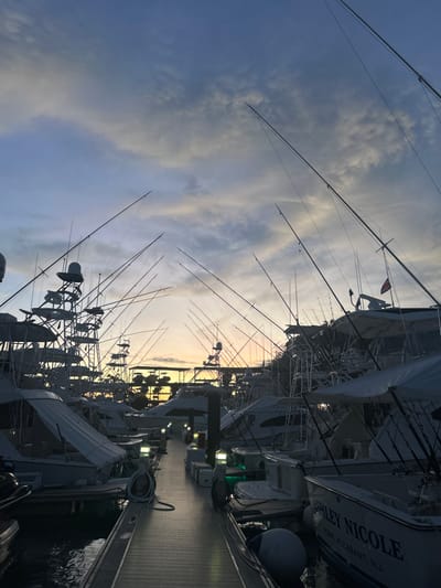 The image shows a marina in Playa Herradura, Costa Rica at sunset. Several large white boats are docked along a wooden pier. The sky is a mix of white, light blue, and dark blue clouds with a hint of orange from the setting sun in the distance. The boats appear to be fishing boats, as many of them have fishing rods mounted on the sides and roofs. One boat is labeled "SHILEY NICOLE, POINT PLEASANT, N.J." The image is shot from the pier, looking down towards the water. The wooden pier is lit by several yellow lights.
