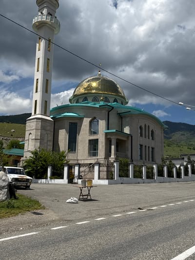 The image shows a mosque with a tall, slender white minaret topped with a golden dome. The mosque building itself is constructed of light-colored stone and features a teal-colored roof. A black power line runs across the frame in front of the mosque and minaret. In the foreground, a white car is parked on the left side of the road. A small, rustic table with two buckets on it is situated in the middle of the paved area. Several white plastic bags are on the ground near the table. The scene is set against a backdrop of rolling green hills and a cloudy sky. The location is identified as Verkhnyaya Teberda, Russia.