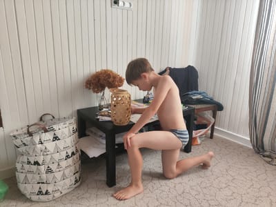 A young boy is kneeling on a patterned carpet next to a black table. He is shirtless and wearing striped underwear. The boy is looking down at something on the table, with his right knee bent and his left leg extended behind him. On the table, there is a wooden decorative lantern, a glass vase with dried flowers, and some papers or magazines. To the left of the table, there is a large fabric storage bag with a teepee pattern. In the background, there is a white wooden paneled wall with a power outlet. To the right, there is a chair with clothes on it and a striped curtain. The scene appears to be indoors, likely a home, with natural light coming from the right. There is no visible text in the image.