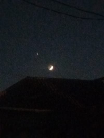 In the dark sky above the city of Sabunchu, Azerbaijan, a sliver of the crescent moon is visible, illuminated by an unseen light source. To the left of the moon, a bright star, possibly Venus, shines. Below the celestial bodies, the silhouette of a dark, sloped roofline is discernible, casting a shadow over the lower portion of the image. The overall scene is one of a quiet, nocturnal landscape.