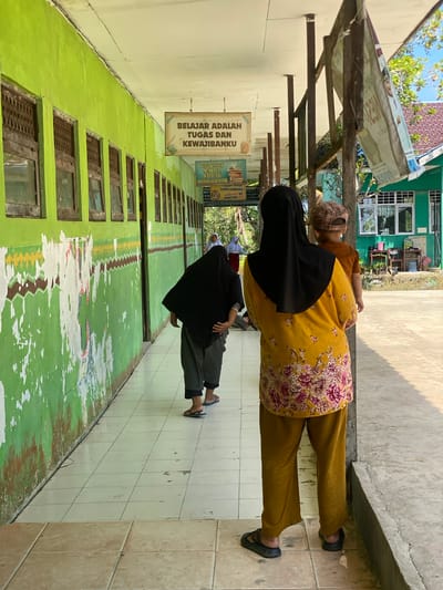 This is an eye-level, medium shot of a walkway along the exterior of a building. The building's wall is painted a bright green and shows signs of wear, with peeling paint and faded murals. Several windows with wooden frames and grated coverings are visible along the wall. Hanging from the ceiling are two signs. The sign closest to the viewer reads "BELAJAR ADALAH TUGAS DAN KEWAJIBANKU" (Learning is my duty and obligation). Further down the walkway, another sign depicts an open book with text.

In the foreground, a woman wearing a black hijab and a yellow shirt with a floral pattern stands with her back to the camera, holding a young child. The child is wearing a checkered cap and an orange shirt. To the left of this woman, another person in a black hijab and grey pants is walking away from the camera. In the background, other individuals, likely students, can be seen further down the walkway. The walkway itself is tiled with light-colored squares, and a concrete gutter runs along the right side of the image. Beyond the walkway and building, a green building with windows and a tiled roof is visible, suggesting this is likely a school in Handil Baru, Indonesia. The scene is illuminated by bright sunlight, casting shadows on the ground.