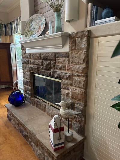 The image depicts a cozy living room scene with a prominent stone fireplace. The fireplace has a white mantelpiece adorned with decorative items, including a large plate with a coral and seahorse design, a blue glass bottle, and a green vase with dried flowers. On the hearth, a blue glass buoy with netting and a nautical-themed wooden buoy are visible. A carved wooden seagull stands to the right of the nautical buoy. The fireplace itself has glass doors that reflect the interior. To the right of the fireplace, a wall with horizontal white paneling is present, partially obscured by a large green plant. The floor is made of polished wood. The overall aesthetic suggests a coastal or beach-themed decor. The image does not provide any specific information about the city of Bradenton, United States.