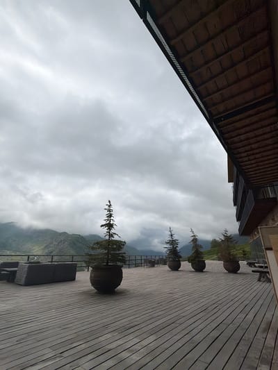 This image depicts an outdoor wooden deck with several potted evergreen trees and outdoor seating. In the background, a mountain range is visible under a cloudy sky. The architecture of a building with a wooden roof and balconies is present on the right side of the frame. The setting appears to be a terrace or viewpoint in the vicinity of Stepantsminda, Georgia, offering a scenic view of the surrounding mountainous landscape. There are no people visible in the image.