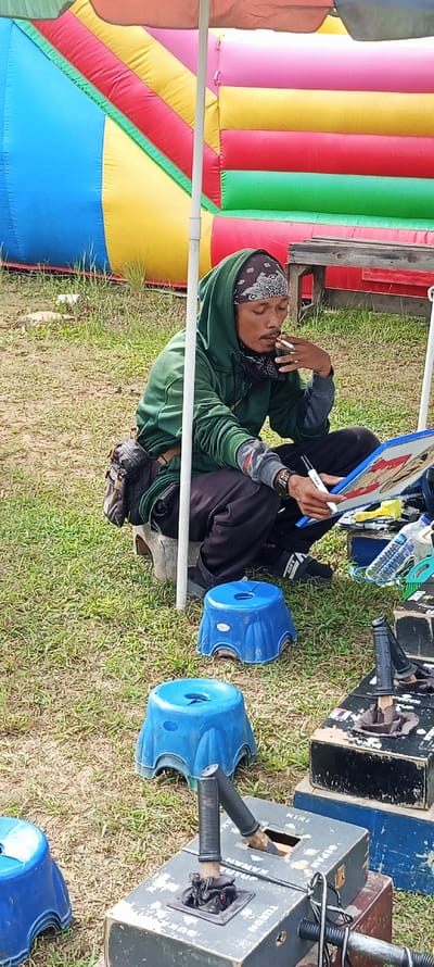 An adult male is observed outdoors in Bandasari, Indonesia, seated on a wooden stool or tree stump. He wears a dark green hooded sweatshirt, a black bandana with a white paisley pattern, dark pants, and black slides. A dark hip bag is slung over his right side, and a watch is on his left wrist. He holds a lit cigarette in his right hand, positioned near his mouth, while his left hand grasps a white marker or pen. He is engaged with a blue-framed board or canvas, which depicts a stylized figure. Behind him, a large, multi-colored inflatable structure (blue, yellow, red, pink, green) is visible. A white pole from an overhead shade structure bisects the frame. On the ground to his right are three blue plastic stools and several dark-colored wooden boxes, some stacked. One box displays Indonesian text, including "KANAN" (right) and "KIRI" (left), and features a joystick-like wooden handle wrapped in black material. A plastic water bottle and a yellow plastic item are also present on the boxes. The ground consists of sparse grass and exposed soil.