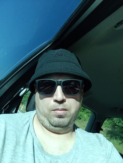 The image shows a man wearing a black bucket hat and sunglasses, sitting in a car. The car's sunroof is open, revealing a bright blue sky. The man is wearing a grey t-shirt. The background outside the car shows green trees and foliage, indicating an outdoor, possibly rural, setting. The car appears to be moving or parked along a road. There is no specific information within the image to confirm the location as Psáře, Czechia.