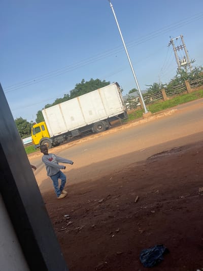 In Kassa, Nigeria, a young boy in a grey hoodie and blue jeans stands on a dirt road, looking at the camera with his arms extended. Behind him, a large white truck is parked on the side of the road. The truck is a box truck with a yellow cab. The road is made of dirt and gravel, and there are trees and a power pole in the background. The sky is blue and clear. There is trash on the ground.