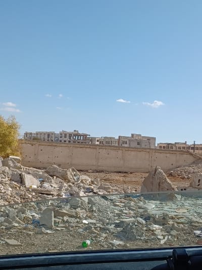 This image depicts a devastated urban landscape in Al-Qadam Municipality, Syria, under a clear blue sky with minimal white cloud cover. The foreground is dominated by a vast expanse of construction rubble, consisting of shattered concrete, broken masonry, rebar fragments, and scattered debris including shards of glass and plastic waste, notably a green bottle on the left. The scene appears to be viewed from inside a vehicle, with a dark dashboard or window frame visible at the bottom. In the midground, a long, tan-colored concrete wall, approximately 2-3 meters high, runs horizontally, displaying numerous impact marks consistent with bullet holes or shrapnel damage. Behind this damaged wall, the ground is barren and dusty. The background features a cluster of multi-story buildings, appearing structurally compromised or unfinished, with numerous open window and door frames suggesting severe damage or incomplete construction. No individuals are present in the scene.