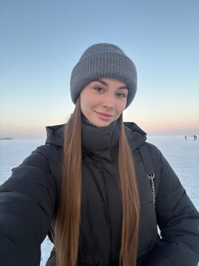 A person in a gray beanie and a black puffer jacket takes a selfie outdoors. The sky is a pale blue with a hint of pink near the horizon. A flat, snowy expanse stretches out behind the person, with a few small figures visible in the distance. The person has long, straight brown hair that falls across their chest. They have light skin, brown eyes, and a slight smile. A silver chain is visible on the jacket.