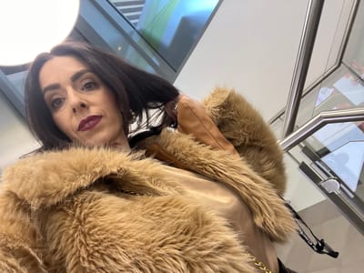 A woman with dark hair and dark lipstick is wearing a brown fur-trimmed coat. A handbag with a gold chain strap is visible. A portion of a modern interior with a glass and metal railing is in the background. The location is possibly within the city of Mensdorf, Luxembourg.