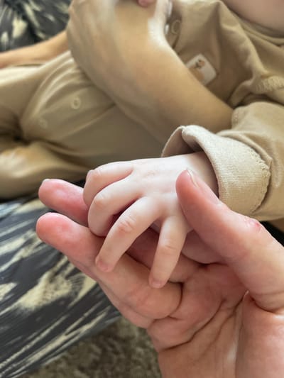 A close-up shot focuses on a baby's hand held by a larger hand, presumably an adult's. The baby is wearing a beige onesie. The adult's hand has visible wrinkles. The background includes striped trousers and a hint of a neutral-toned carpet. The setting is not discernible enough to determine the location in Crokhovo, Russia or any city.